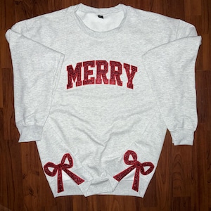 Pode incluir: Uma camisa de moletom cinza com a palavra "MERRY" em glitter vermelho na frente. As mangas têm laços de glitter vermelho nas mangas.