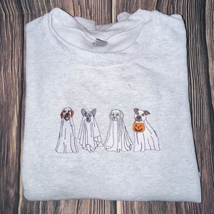 以下が含まれることがあります： ハロウィンのために幽霊に扮した4匹の犬が刺繍されたグレーのスウェットシャツ。3匹の犬は伝統的な幽霊の衣装を着ており、4匹目の犬はジャック・オ・ランタンを持っています。
