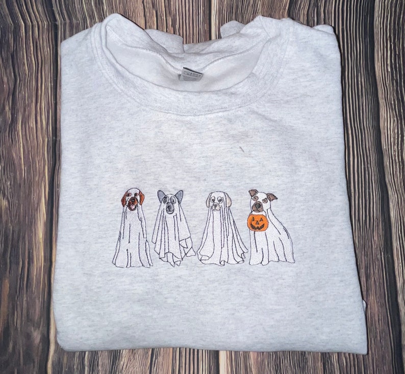 以下が含まれることがあります： ハロウィンのために幽霊に扮した4匹の犬が刺繍されたグレーのスウェットシャツ。3匹の犬は伝統的な幽霊の衣装を着ており、4匹目の犬はジャック・オ・ランタンを持っています。