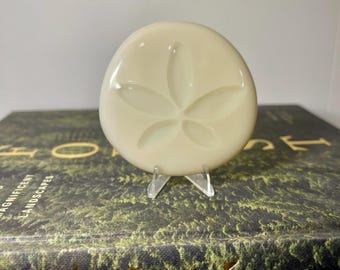 May include: A cream-colored, round sand dollar with a five-petal design, displayed on a clear plastic stand. The sand dollar is smooth and glossy, and the stand is positioned on a book with gold lettering and a green and brown cover.