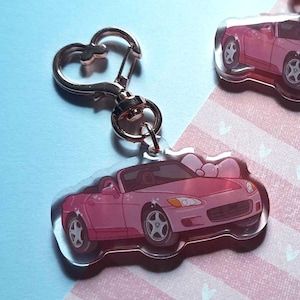 May include: Three pink car-shaped keychains with rose gold heart-shaped clasps. The cars have a bow design on the front and are set against a light blue and pink striped background. The keychains are made of clear acrylic.