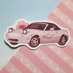 May include: A sticker depicting a light pink car with a black roof and cherry blossom details. The car has pop-up headlights and a floral design. The sticker is on a pink and blue background.