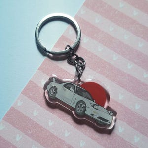 Può includere: Un portachiavi in acrilico trasparente con un design di auto sportiva bianca e un sole rosso. Il portachiavi ha un anello e una catena in metallo argentato. Lo sfondo è un motivo a strisce rosa e bianche.