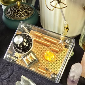 Peut inclure: Une boîte en plastique transparente contenant un circuit imprimé doré. Le circuit imprimé comporte un bouton avec des chiffres de 0 à 100, des bobines de cuivre et un petit cristal jaune. La boîte est ouverte d'un côté, révélant les composants.