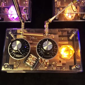 May include: A clear plastic box with two black knobs labeled 10 to 100, a copper coil, and a small, glowing orange crystal. The box is on a black surface.