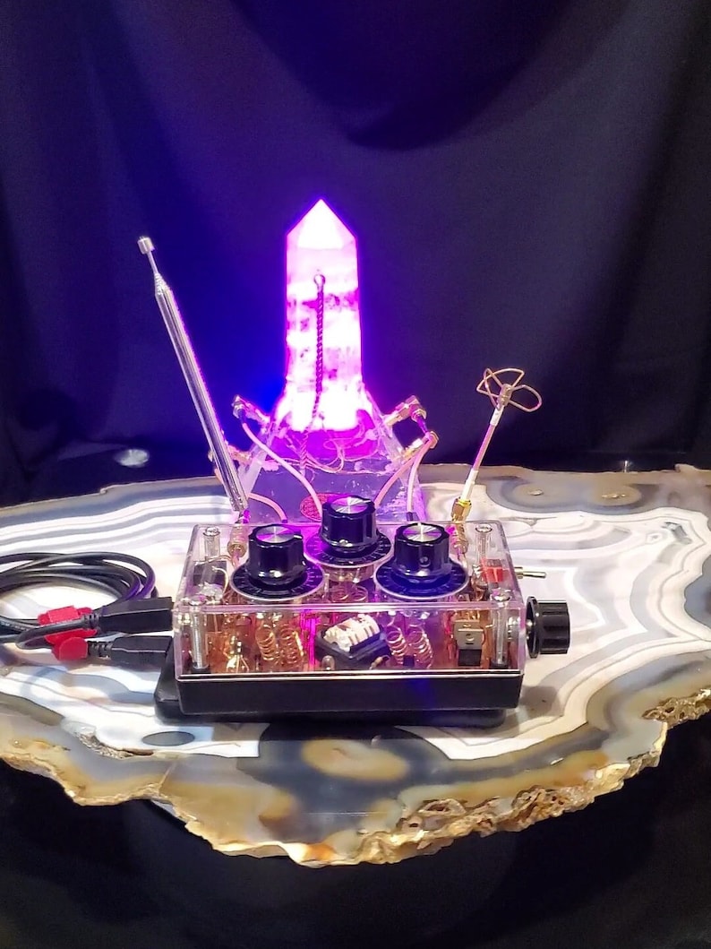 May include: A clear plastic box with a black base containing a circuit board with three knobs and a purple crystal on top. The crystal is illuminated with a purple light. The box is sitting on a large, gray and brown agate stone.