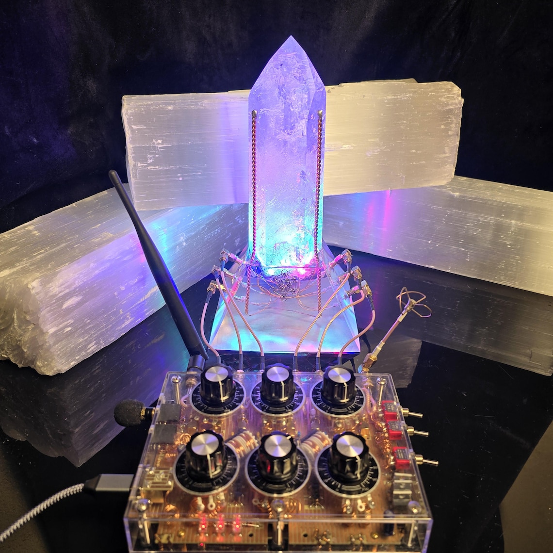 May include: A clear quartz crystal illuminated with blue and purple light, mounted on a clear acrylic base. The base is connected to a control panel with knobs and wires. Two large white selenite slabs are in the background.
