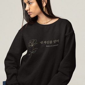 Puede incluir: Sudadera negra con un contorno dorado de una rosa y el texto "네 자신을 믿어", que se traduce como "Cree en ti mismo" en español.