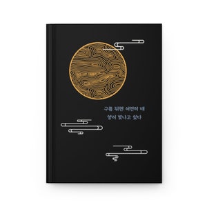以下が含まれることがあります： 黒い本の表紙に、金の円形のデザインと韓国語で「구름 뒤편 햇살이 떼  향이 빛나고 있다」というテキストが書かれている。