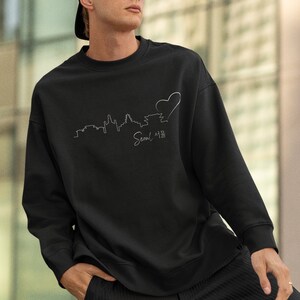 May include: Black sweatshirt with a white outline of the Seoul skyline and a heart. The text "Seoul 서울" is written below the skyline.