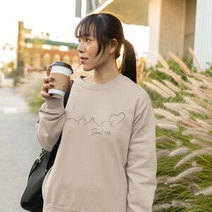 May include: A woman wearing a beige sweatshirt with a black outline of the Seoul skyline and a heart, with the word "Seoul 서울" written below. She is holding a brown paper cup and looking to the right.