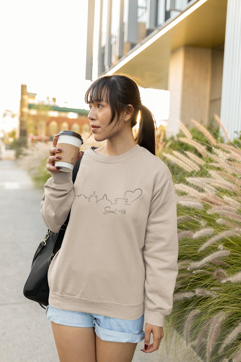 May include: A woman wearing a beige sweatshirt with a black outline of the Seoul skyline and a heart, with the word "Seoul 서울" written below. She is holding a brown paper cup and looking to the right.