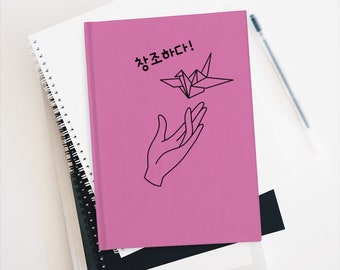 May include: A pink notebook with a black line drawing of a hand reaching out to a paper crane. The text "잘 조하다!" is written in black on the notebook.