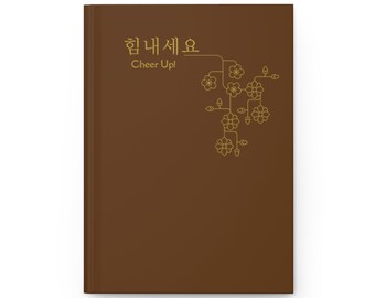 May include: A brown notebook with the Korean text "힘내세요" and the English text "Cheer Up!" in gold lettering. A stylized gold floral design is on the right side of the notebook.