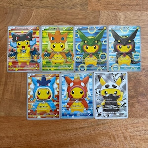 May include: A collection of seven Pokémon trading cards featuring Pikachu wearing various character-themed hoods, including Charizard, Rayquaza, Umbreon, Gyarados, Magikarp, and Skull-themed outfits, with Japanese text printed on each card surface.