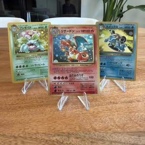 May include: Three Japanese Pokemon trading cards displayed on clear plastic stands. The left card is green featuring Venusaur, the center card is red featuring Charizard, and the right card is blue featuring Blastoise. Each card displays character art, HP values, and Japanese text.