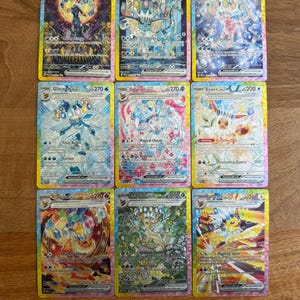 May include: A collection of nine rectangular Pokémon trading cards arranged in a three-by-three grid. Each card features a unique character illustration with holographic foil details, text, and game statistics, including Umbreon, Vaporeon, Espeon, Glaceon, Sylveon, Eevee, Flareon, Leafeon, and Jolteon.