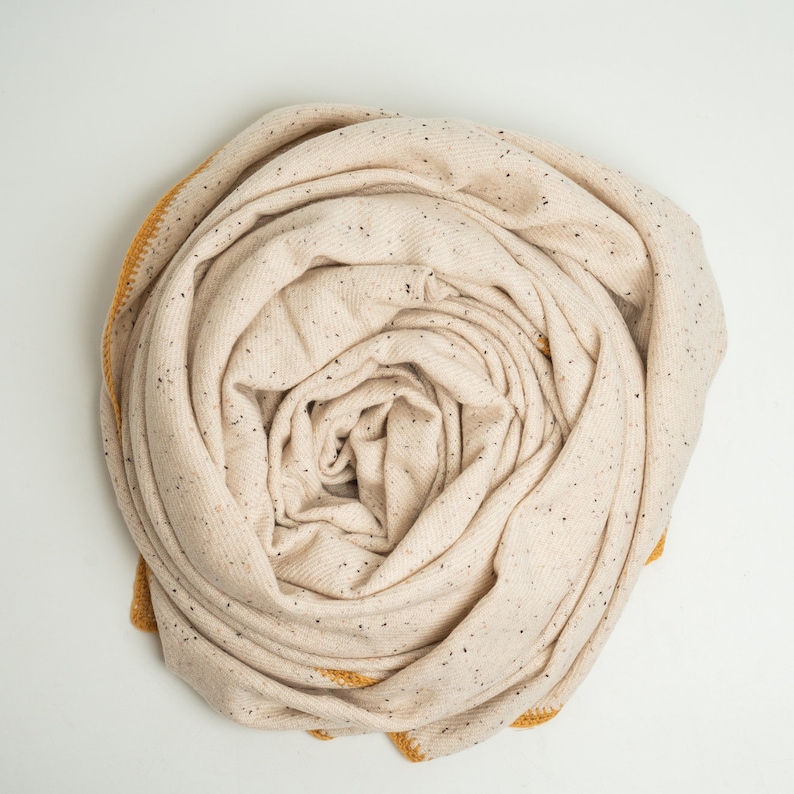 K&ouml;nnte beinhalten: Gerollter beigefarbener Schal mit schwarzen Sprenkeln und einem senfgelben Rand. Der Schal besteht aus einem weichen, gewebten Material und wird auf wei&szlig;em Hintergrund pr&auml;sentiert. Der Schal ist spiralf&ouml;rmig aufgerollt.
