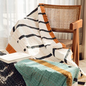 K&ouml;nnte beinhalten: Eine wei&szlig;e, schwarze und orange gestreifte Wolldecke mit einem gesteppten Muster liegt &uuml;ber einem Korbstuhl. Die Decke ist gefaltet und hat ein Buch darauf.