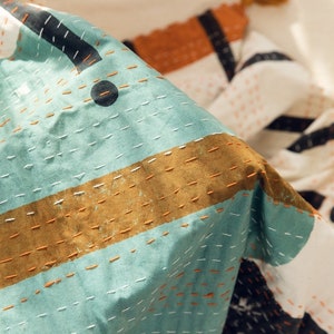 K&ouml;nnte beinhalten: Nahaufnahme einer tealblauen und braunen gemusterten Steppdecke mit wei&szlig;en N&auml;hten. Die Steppdecke ist aus einem weichen, strukturierten Stoff gefertigt und hat ein geometrisches Design.