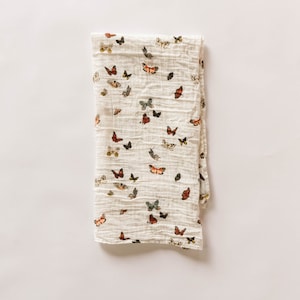 May include: A folded, cream-colored muslin blanket featuring a repeating pattern of colorful butterflies. The butterflies are in shades of red, orange, black, and gray. The blanket has a crinkled texture and is set against a white background.