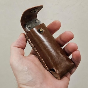 May include: A brown leather sheath with visible stitching along the edges and a metal snap button closure. The sheath contains a silver-colored multi-tool with exposed metal components visible at the top opening of the leather case.