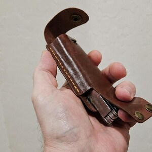 May include: A brown leather sheath with visible yellow stitching and two brass-colored metal snap fasteners. The sheath is designed to hold a multi-tool, which is partially visible inside the leather casing, featuring a curved top flap.