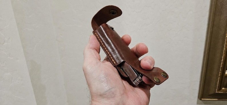 May include: A brown leather sheath with visible yellow stitching and two brass-colored metal snap fasteners. The sheath is designed to hold a multi-tool, which is partially visible inside the leather casing, featuring a curved top flap.