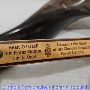 Puede incluir: Una correa de cuero marr&oacute;n con una incrustaci&oacute;n rectangular de color tostado claro con el texto grabado: Hear, O Israel! יהוה is our Elohim, יהוה is One! Blessed is the Name of His Glorious Kingdom For all Eternity. La pieza est&aacute; fijada con remaches de lat&oacute;n.