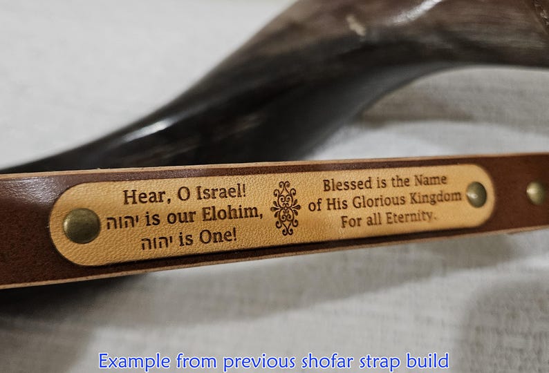 Puede incluir: Una correa de cuero marr&oacute;n con una incrustaci&oacute;n rectangular de color tostado claro con el texto grabado: Hear, O Israel! יהוה is our Elohim, יהוה is One! Blessed is the Name of His Glorious Kingdom For all Eternity. La pieza est&aacute; fijada con remaches de lat&oacute;n.
