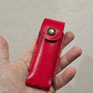 May include: A bright red leather sheath featuring visible stitching along the edges and a single brass-colored metal snap button closure on the flap. The rectangular pouch is designed to hold a small tool or accessory securely in place.
