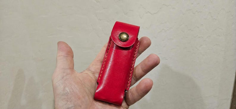 May include: A bright red leather sheath featuring visible stitching along the edges and a single brass-colored metal snap button closure on the flap. The rectangular pouch is designed to hold a small tool or accessory securely in place.
