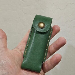 May include: A forest green leather sheath featuring a flap closure secured with a single brass-toned metal snap button. The edges of the sheath are joined with visible, thick, light-colored stitching along the sides, creating a structured pouch.