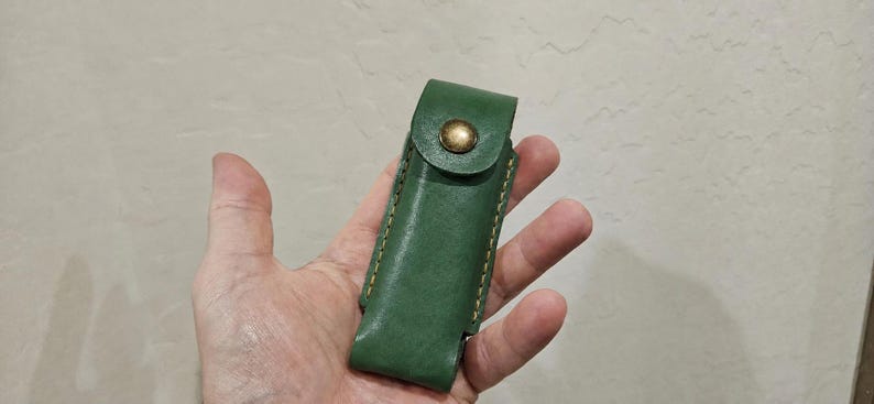 May include: A forest green leather sheath featuring a flap closure secured with a single brass-toned metal snap button. The edges of the sheath are joined with visible, thick, light-colored stitching along the sides, creating a structured pouch.