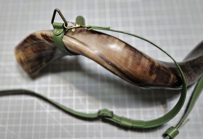 Puede incluir: Un cuerno marr&oacute;n pulido y curvado con un patr&oacute;n de veteado natural, equipado con una correa de cuero verde salvia. La correa incluye un anillo triangular de metal en tono lat&oacute;n, un mosquet&oacute;n y una hebilla ajustable para una fijaci&oacute;n segura.