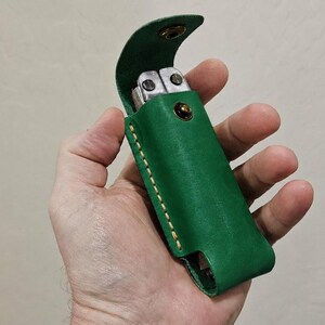 May include: A vibrant green leather sheath designed to hold a multi-tool. The sheath features a flap with a metallic snap closure, visible light-colored stitching along the edges, and an open bottom design to accommodate the tool handle.