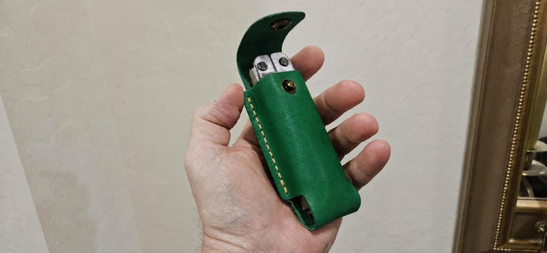 May include: A vibrant green leather sheath designed to hold a multi-tool. The sheath features a flap with a metallic snap closure, visible light-colored stitching along the edges, and an open bottom design to accommodate the tool handle.