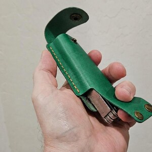 May include: A vibrant green leather sheath with visible tan stitching and two brass-colored metal snap fasteners. The sheath is designed to hold a multi-tool, which is partially visible inside the open structure of the leather casing.
