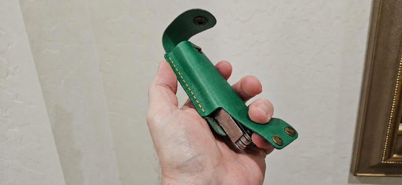 May include: A vibrant green leather sheath with visible tan stitching and two brass-colored metal snap fasteners. The sheath is designed to hold a multi-tool, which is partially visible inside the open structure of the leather casing.