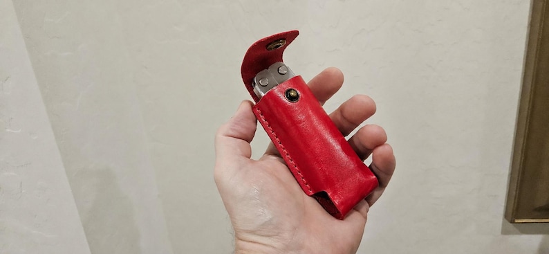 May include: A bright red leather sheath with a snap button closure, designed to hold a multi-tool. The sheath features visible stitching along the edges and a flap that secures the metallic tool inside, which is partially visible at the top.