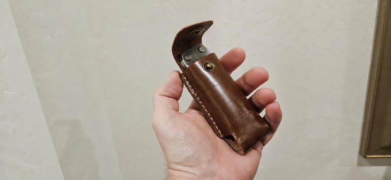 May include: A brown leather sheath with visible stitching along the edges and a metal snap button closure. The sheath contains a silver-colored multi-tool with exposed metal components visible at the top opening of the leather case.