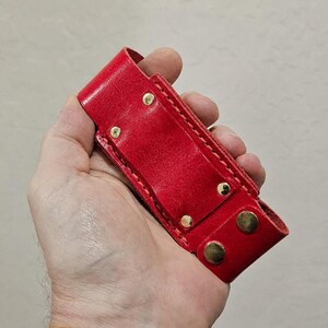 May include: A bright red leather sheath featuring a rectangular body with visible stitching along the edges. The sheath is secured with four metallic gold-toned rivets and two matching gold-toned snap fasteners at the bottom closure flap.