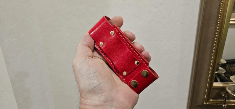 May include: A bright red leather sheath featuring a rectangular body with visible stitching along the edges. The sheath is secured with four metallic gold-toned rivets and two matching gold-toned snap fasteners at the bottom closure flap.