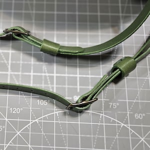 Puede incluir: Dos correas ajustables para bolso en color verde oliva con hebillas met&aacute;licas oscuras y trabillas deslizantes. Las correas presentan m&uacute;ltiples orificios preperforados para ajustar la longitud, con una textura lisa y bordes acabados.