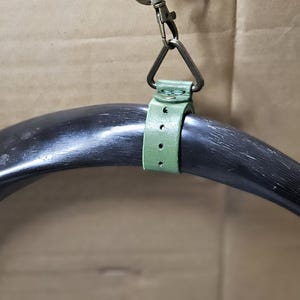 Puede incluir: Un cuerno negro pulido y curvado suspendido por una correa de cuero verde salvia. La correa presenta una serie de agujeros perforados y est&aacute; asegurada con un remache de metal color lat&oacute;n. Un anillo de metal triangular conecta la correa a un mosquet&oacute;n.
