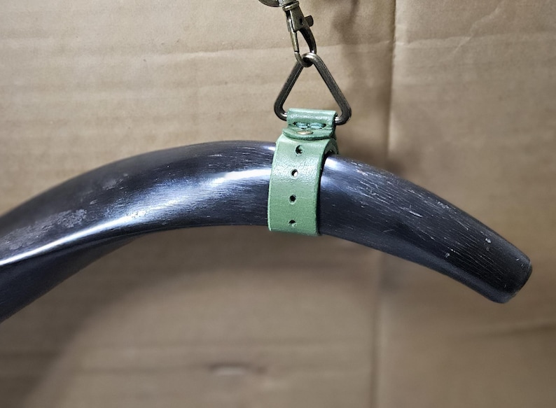 Puede incluir: Un cuerno negro pulido y curvado suspendido por una correa de cuero verde salvia. La correa presenta una serie de agujeros perforados y est&aacute; asegurada con un remache de metal color lat&oacute;n. Un anillo de metal triangular conecta la correa a un mosquet&oacute;n.
