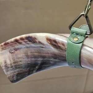 Puede incluir: Un cuerno animal pulido y curvado con vetas naturales en marr&oacute;n y crema, asegurado por una correa de cuero verde salvia. La correa cuenta con un remache de metal color lat&oacute;n y est&aacute; unida a un anillo triangular de metal con un clip giratorio.