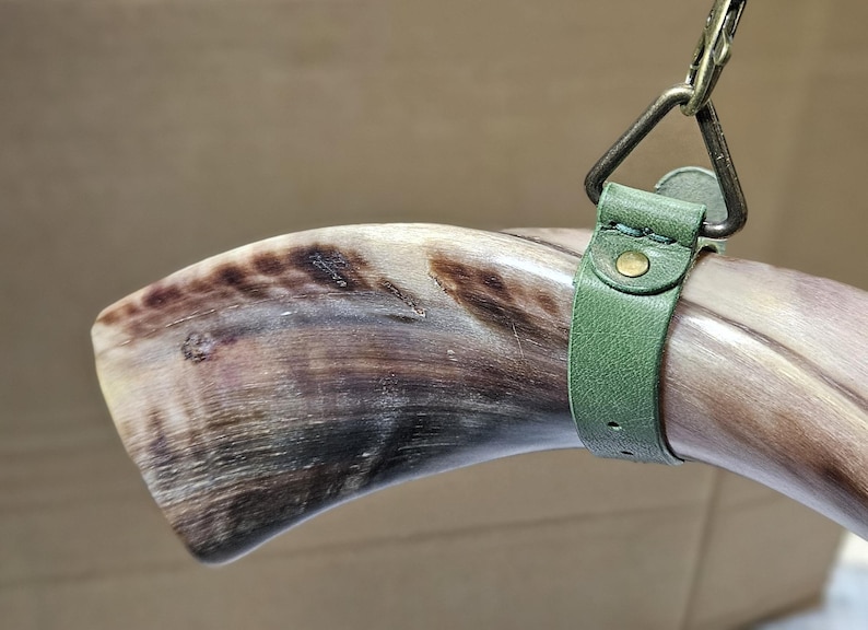 Puede incluir: Un cuerno animal pulido y curvado con vetas naturales en marr&oacute;n y crema, asegurado por una correa de cuero verde salvia. La correa cuenta con un remache de metal color lat&oacute;n y est&aacute; unida a un anillo triangular de metal con un clip giratorio.