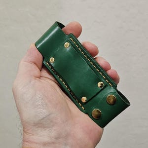 May include: A forest green leather sheath featuring gold-toned metal rivets and snap fasteners. The rectangular holder has visible yellow stitching along the edges and a belt loop attachment on the back, designed for carrying a small tool or knife.