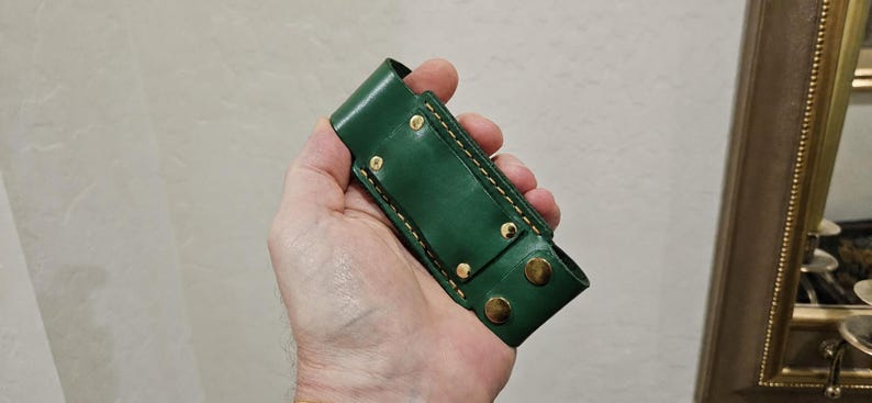 May include: A forest green leather sheath featuring gold-toned metal rivets and snap fasteners. The rectangular holder has visible yellow stitching along the edges and a belt loop attachment on the back, designed for carrying a small tool or knife.